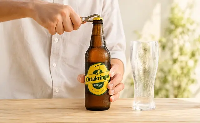 Person öffnet eine Flasche Ottakringer Helles mit einem Flaschenöffner neben ein
