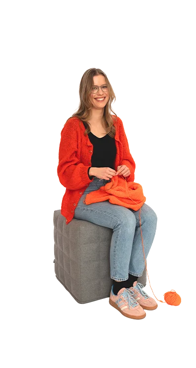 Polina Yaroshchuk, BA MA sitzt mit oranger Strickjacke und Wolle auf einem Hocke